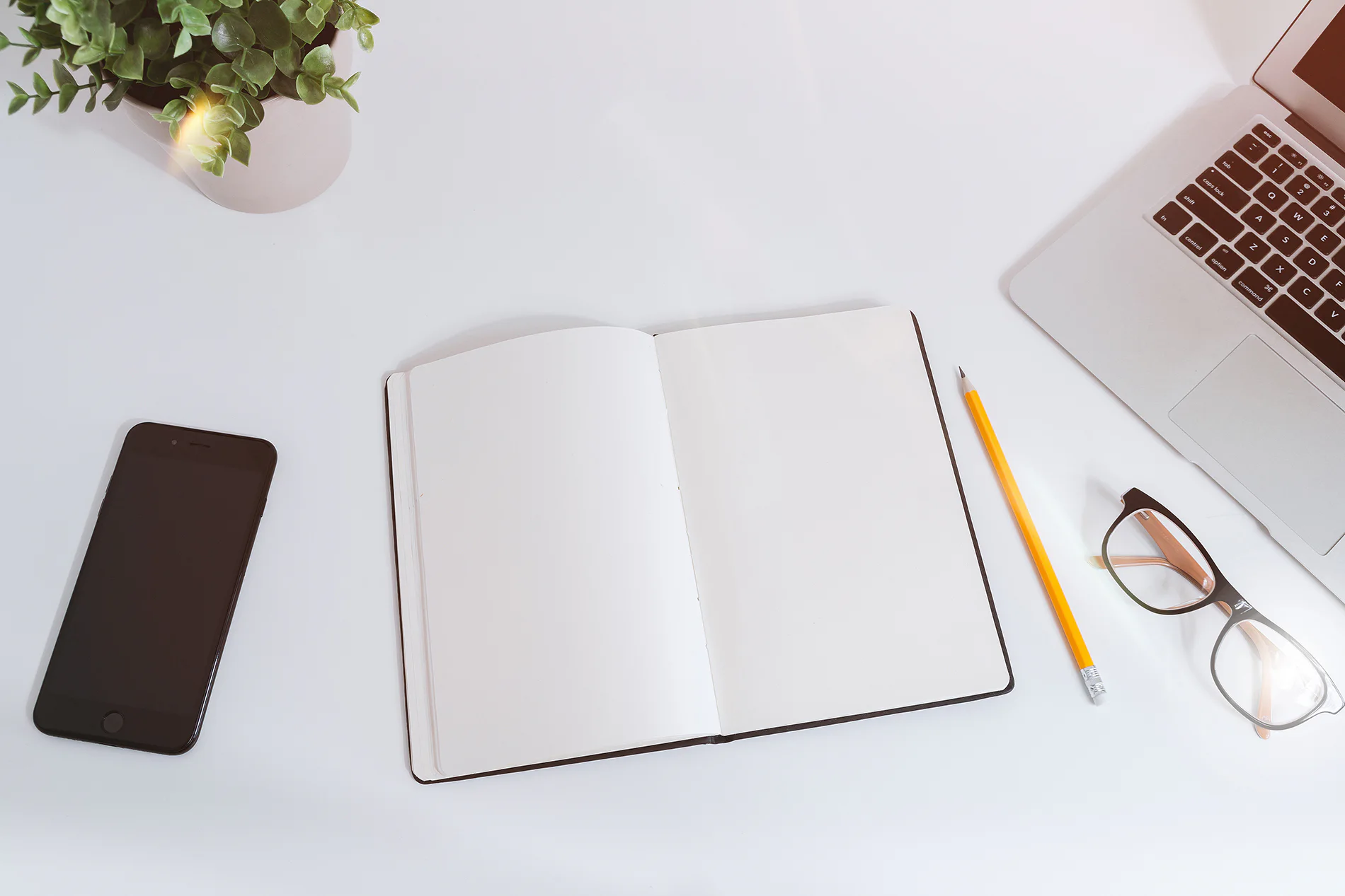 Bright white desk with a notebook, pencil, glasses, phone and laptop
